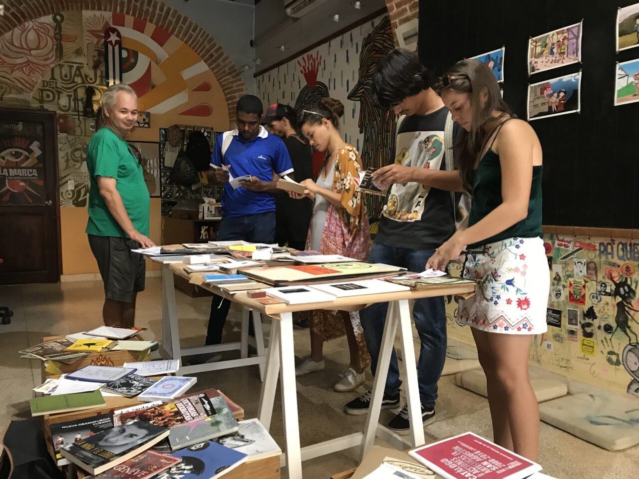 Varias personas hojean publicaciones sobre una mesa de caballete, en un espacio con las paredes cubiertas de carteles.